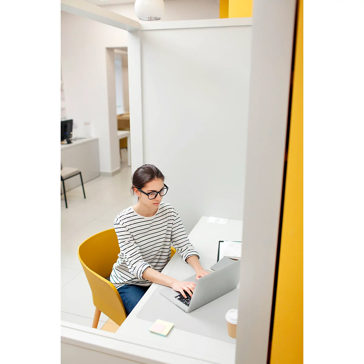 Une femme avec des lunettes est assise à une table devant un ordinateur portable. Elle porte une chemise rayée et un jean, sa chaise est jaune.