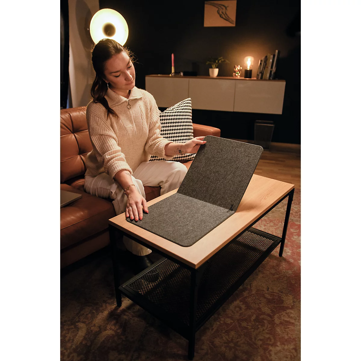 Jeune femme assise sur un canapé, plaçant un coussin en feutre gris sur une table en bois. En arrière-plan : meubles, décorations.