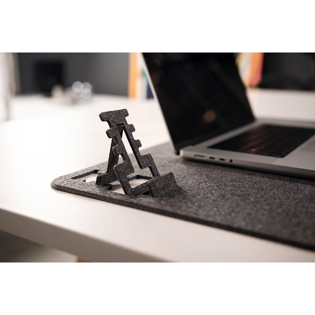 Un support d'ordinateur portable en feutre gris est posé à côté d'un ordinateur portable sur un tapis en feutre gris. L'ordinateur portable est ouvert.