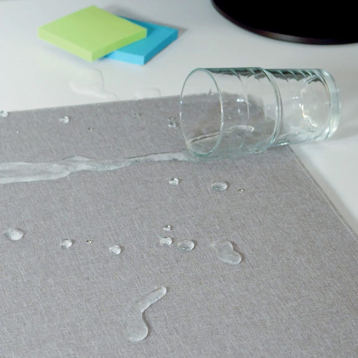 Un verre d'eau renversé sur un tapis de bureau gris, à côté de blocs-notes.
