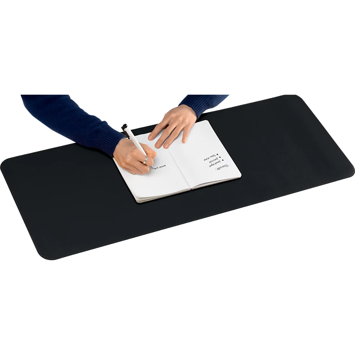 Une personne écrit dans un carnet sur un tapis de bureau noir. Le stylo est dans la main, le carnet est ouvert.