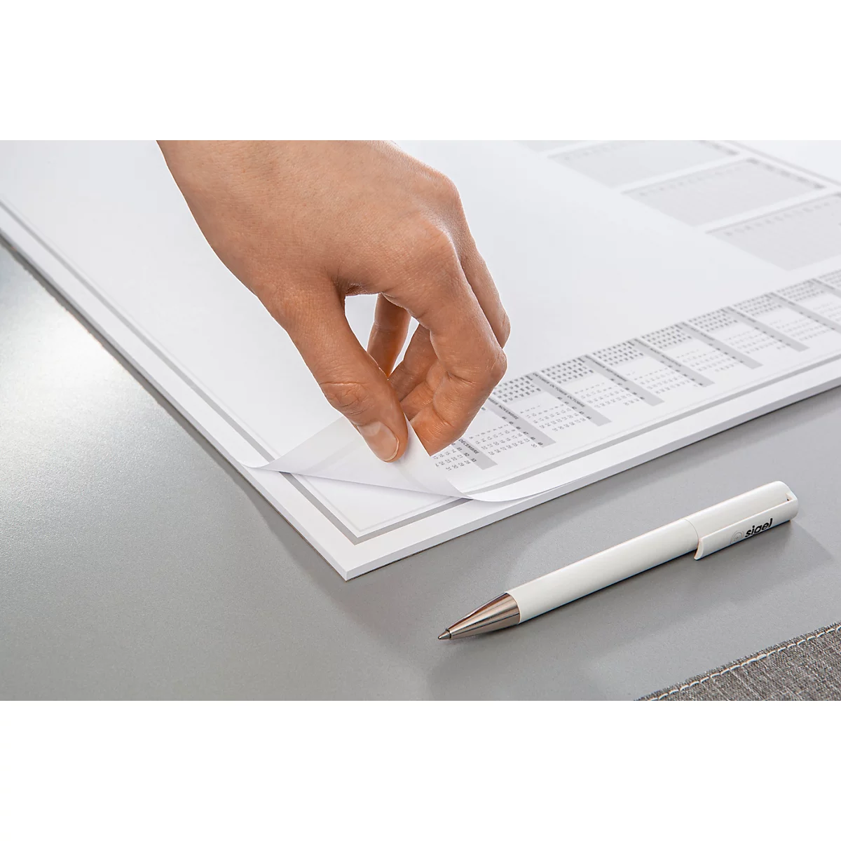 La main soulève une feuille de papier. En dessous, il y a un tableau avec des lignes fines, à côté duquel se trouve un stylo blanc.