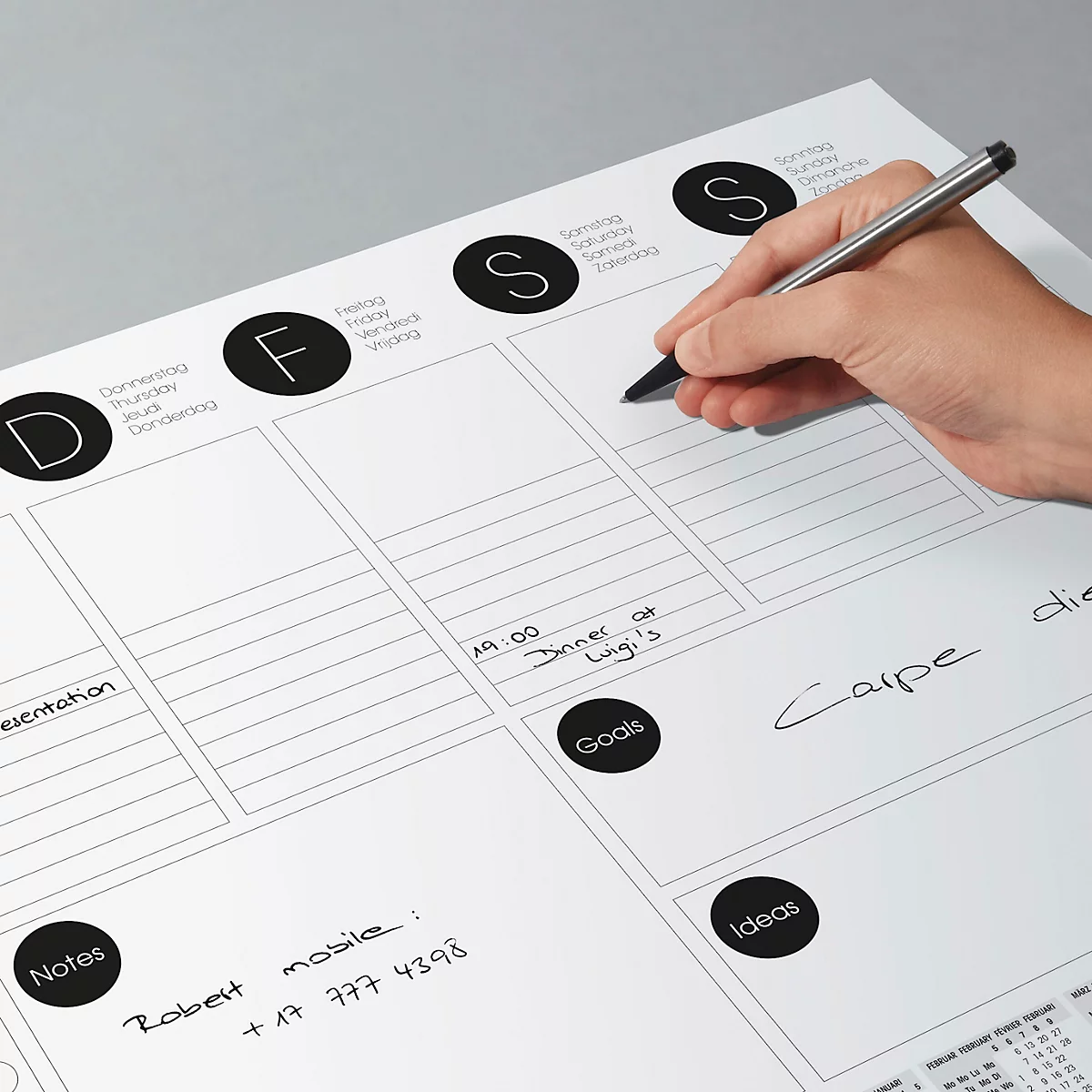 Une personne écrit avec un stylo dans un planificateur hebdomadaire blanc grand format. Le planificateur a des cercles noirs avec des lettres pour les jours de la semaine.