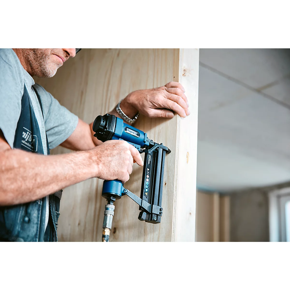 Een timmerman gebruikt een blauwe pneumatische tacker. Hij bevestigt hout aan een muur. Grijze achtergrond.