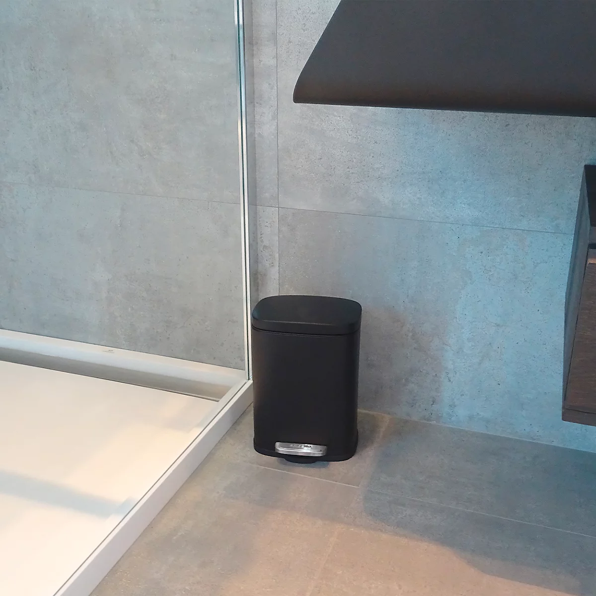 Poubelle noire dans une salle de bain. Elle se trouve devant un mur gris, à côté d'une douche.