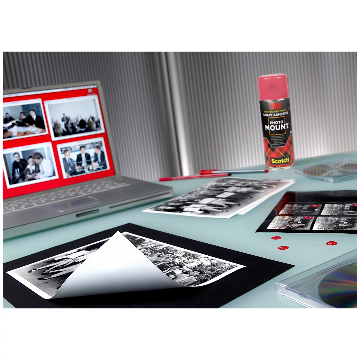 A workstation with a laptop, photos, adhesive spray, and CDs. The laptop displays images on a red interface.