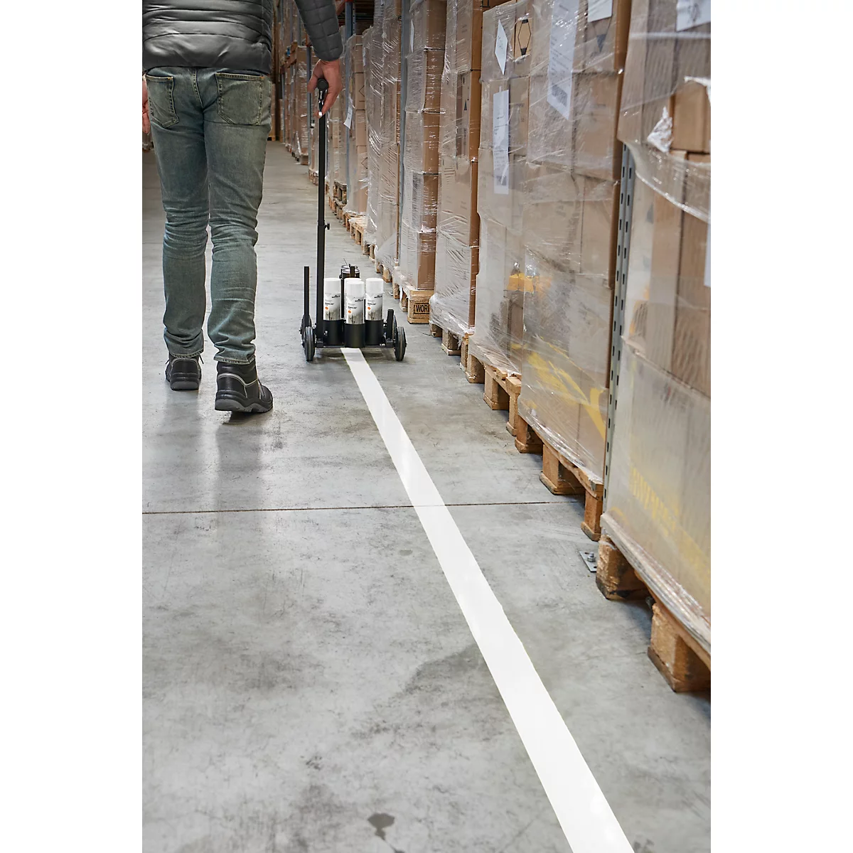 Une personne tire un chariot de marquage, traçant une ligne blanche sur le sol en béton, dans l'entrepôt. Des palettes de marchandises sont en arrière-plan.