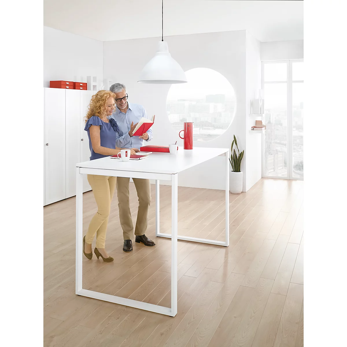 Two people stand at a white table, looking at a red book. Bright interior with a large window.