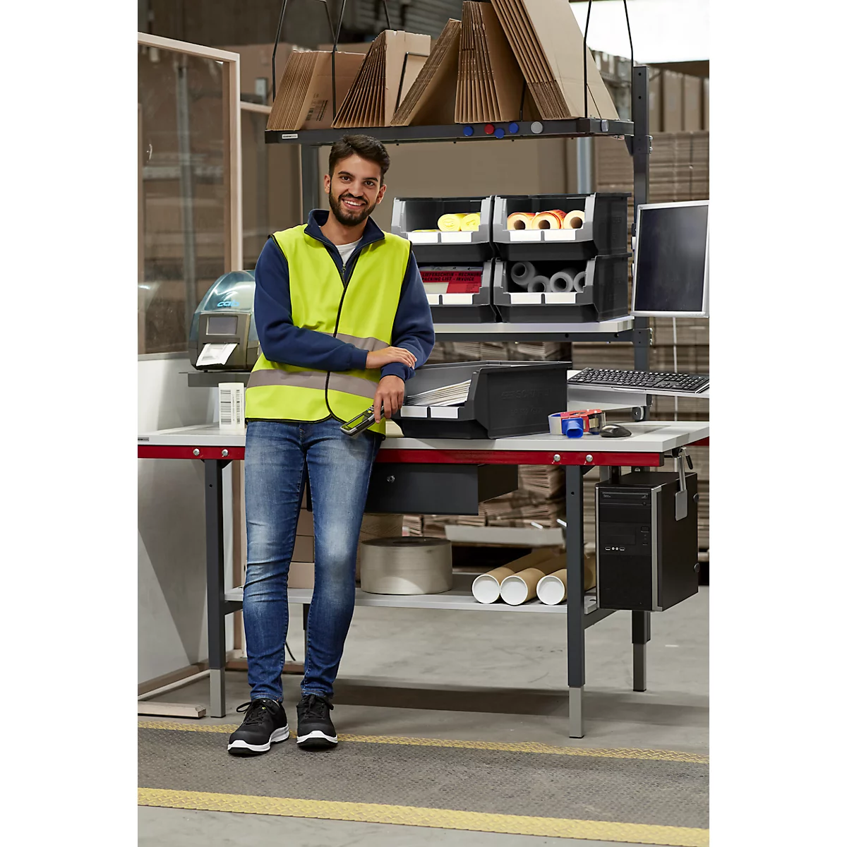 Un homme en gilet jaune s'appuie sur une table dans un entrepôt, souriant à la caméra. Des étagères et des ordinateurs sont derrière lui.