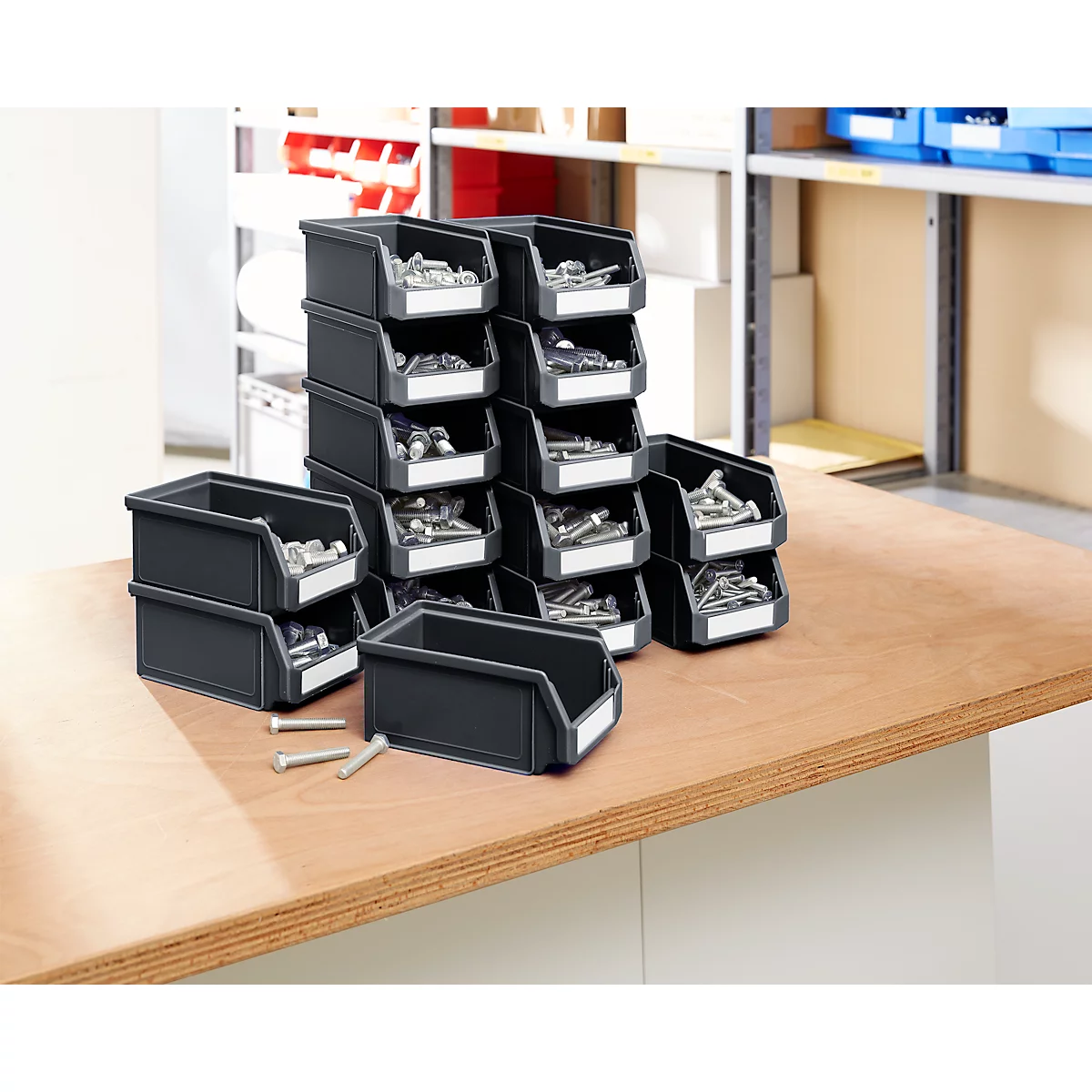 Schwarze Kunststoffbehälter, gefüllt mit Schrauben und Muttern, auf einer Holzoberfläche gestapelt. Einige Behälter haben leere, weiße Etiketten.