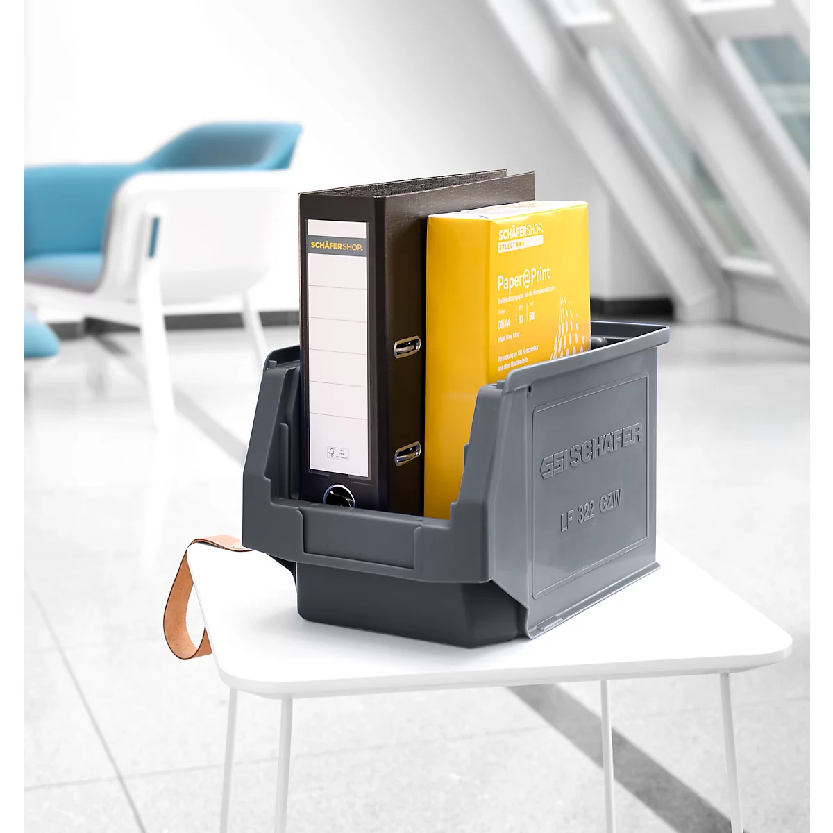 Graue Aufbewahrungsbox mit Ordnern und Papier auf einem weißen Tisch. Im Hintergrund sind Stühle.