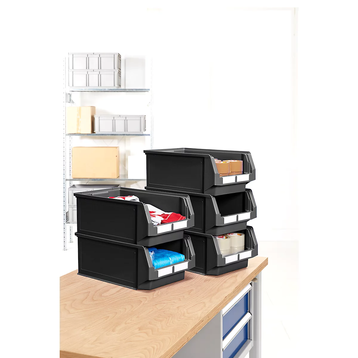 Schwarze Stapelboxen mit Inhalten wie Kleidung und Kartons. Auf einem Holztisch platziert, weitere Boxen im Hintergrund.