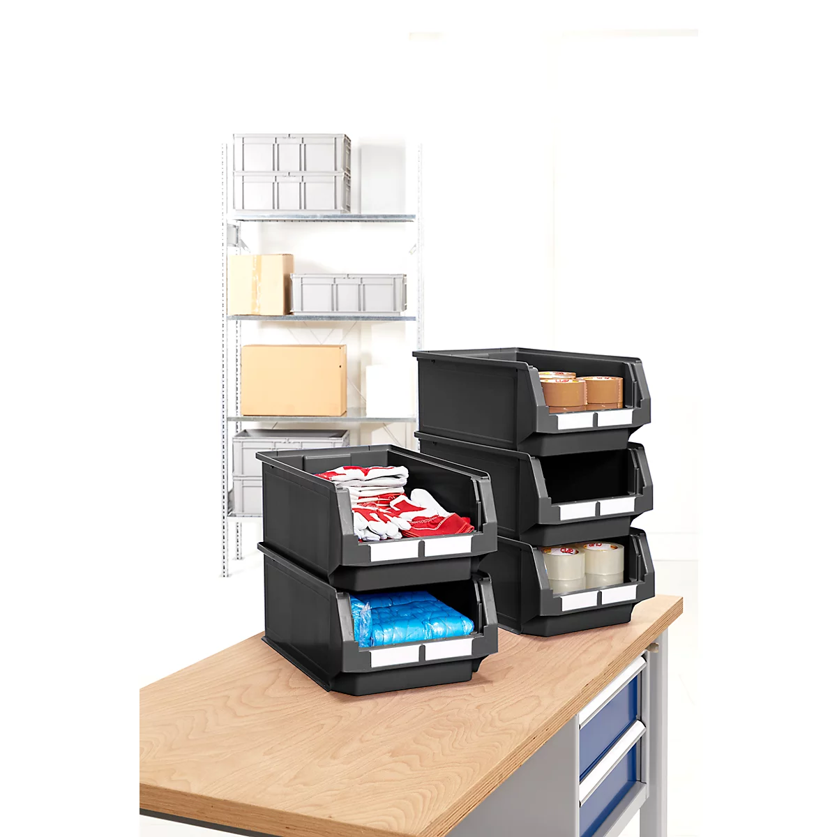 Schwarze Stapelboxen auf einem Holztisch. Gefüllt mit Handschuhen, Folien, Klebeband und Waren. Im Hintergrund ein Regal mit Boxen.