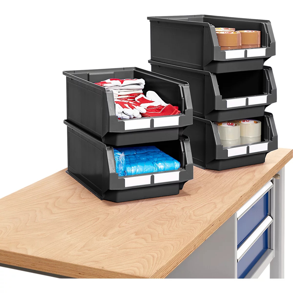 Schwarze Stapelboxen auf einer Werkbank, gefüllt mit Material. Handschuhe, Papierrollen und verpackte Güter sind sichtbar.