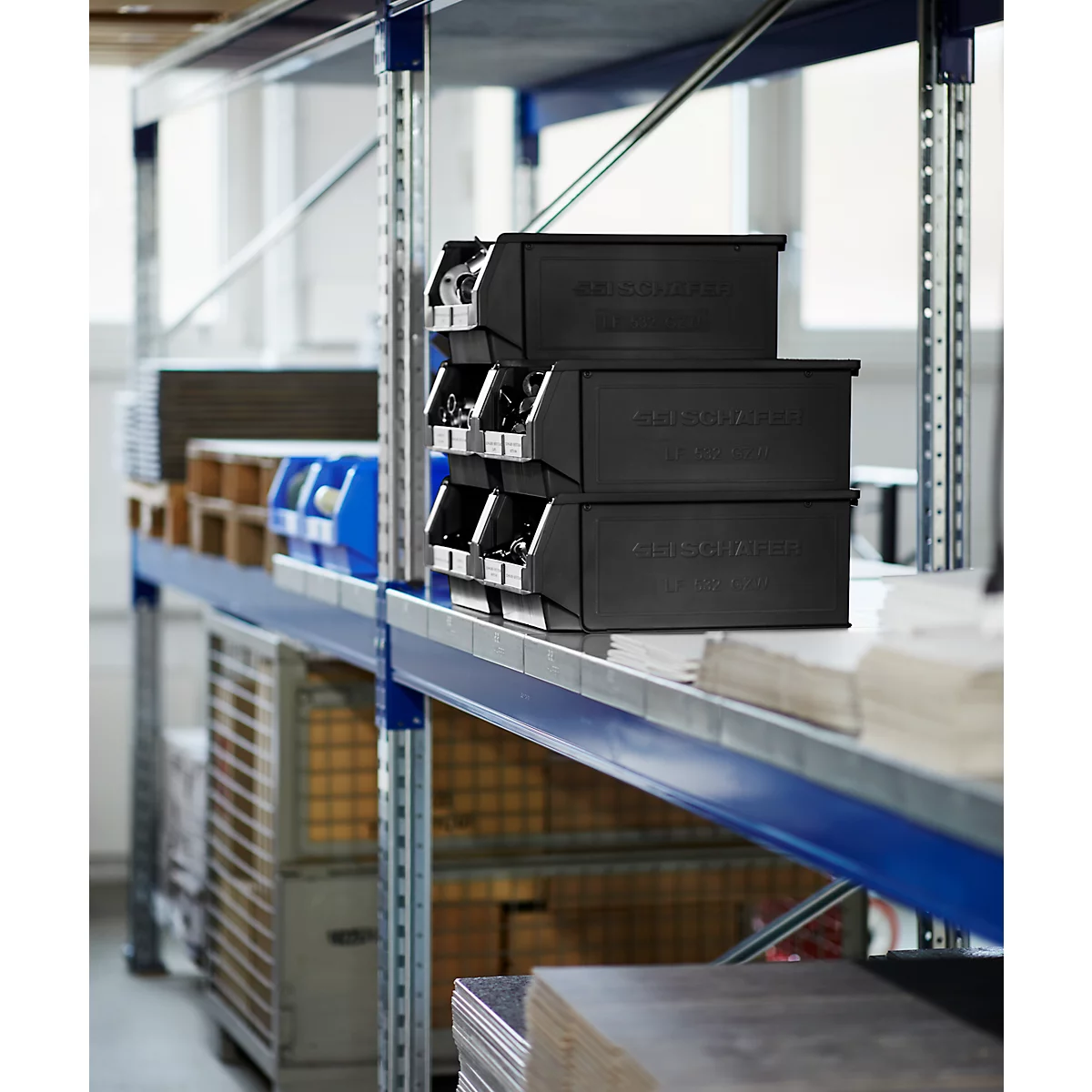 Schwarze Stapelboxen auf einem Metallregal, dahinter weitere Boxen, Kisten und Paletten.