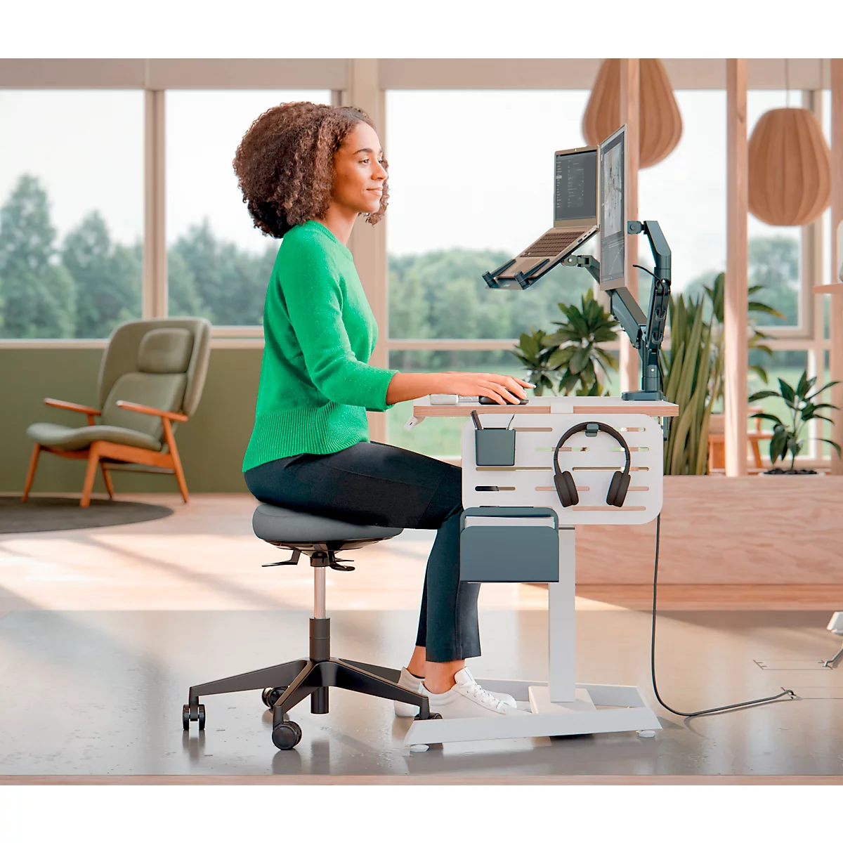 Een vrouw werkt aan een ergonomisch sta-bureau met laptop, monitor en koptelefoon. Ze zit op een bureaustoel. Licht, vriendelijk kantoor.