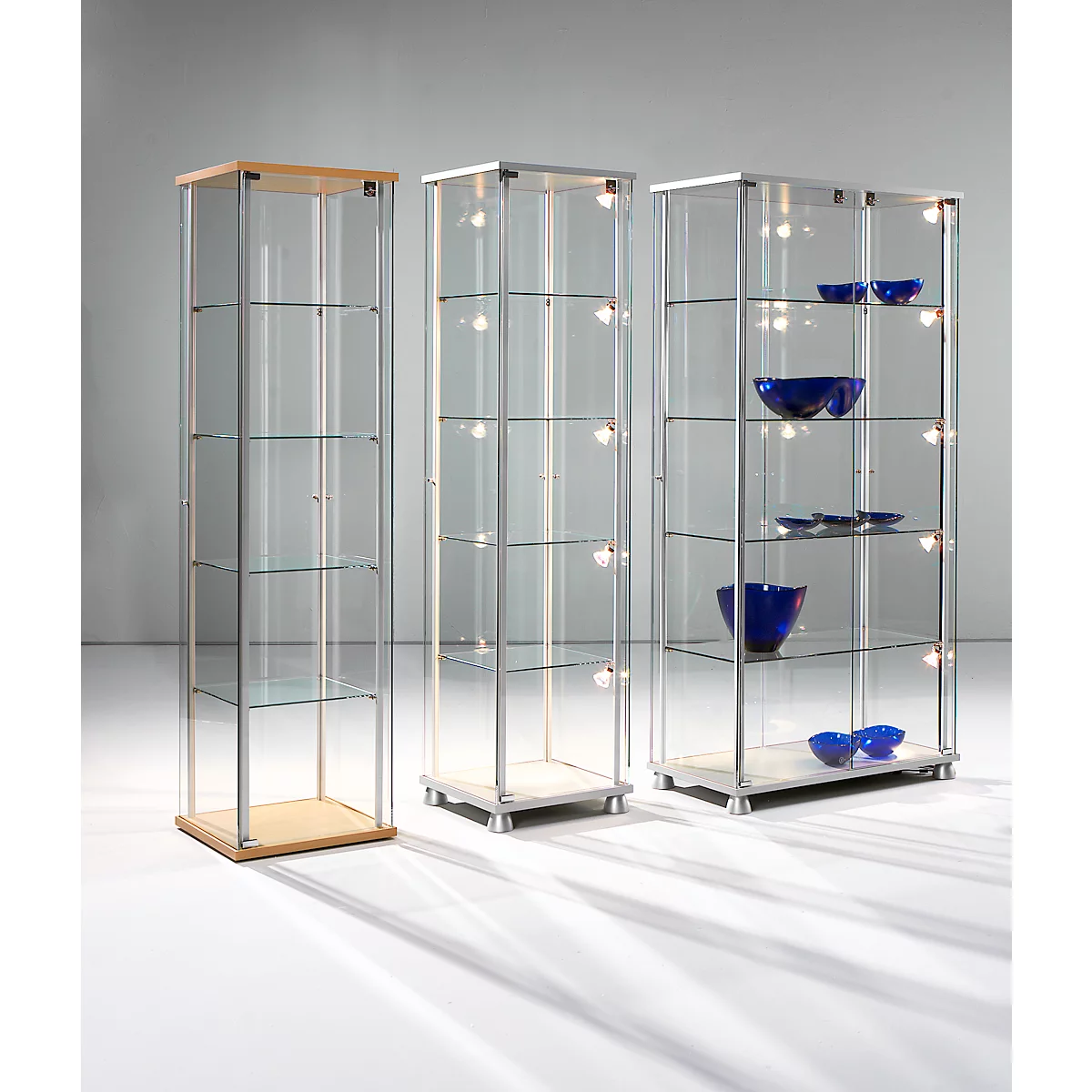 Drie moderne glazen vitrinekasten in verschillende breedtes en ontwerpen tegen een neutrale wand.