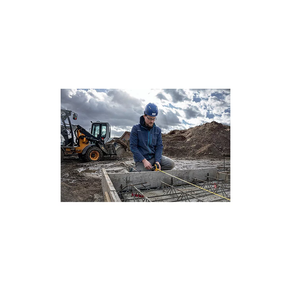 Een bouwvakker knielt bij de fundering van een bouwplaats en meet met een geel meetlint. Op de achtergrond een graafmachine en donkere wolken.