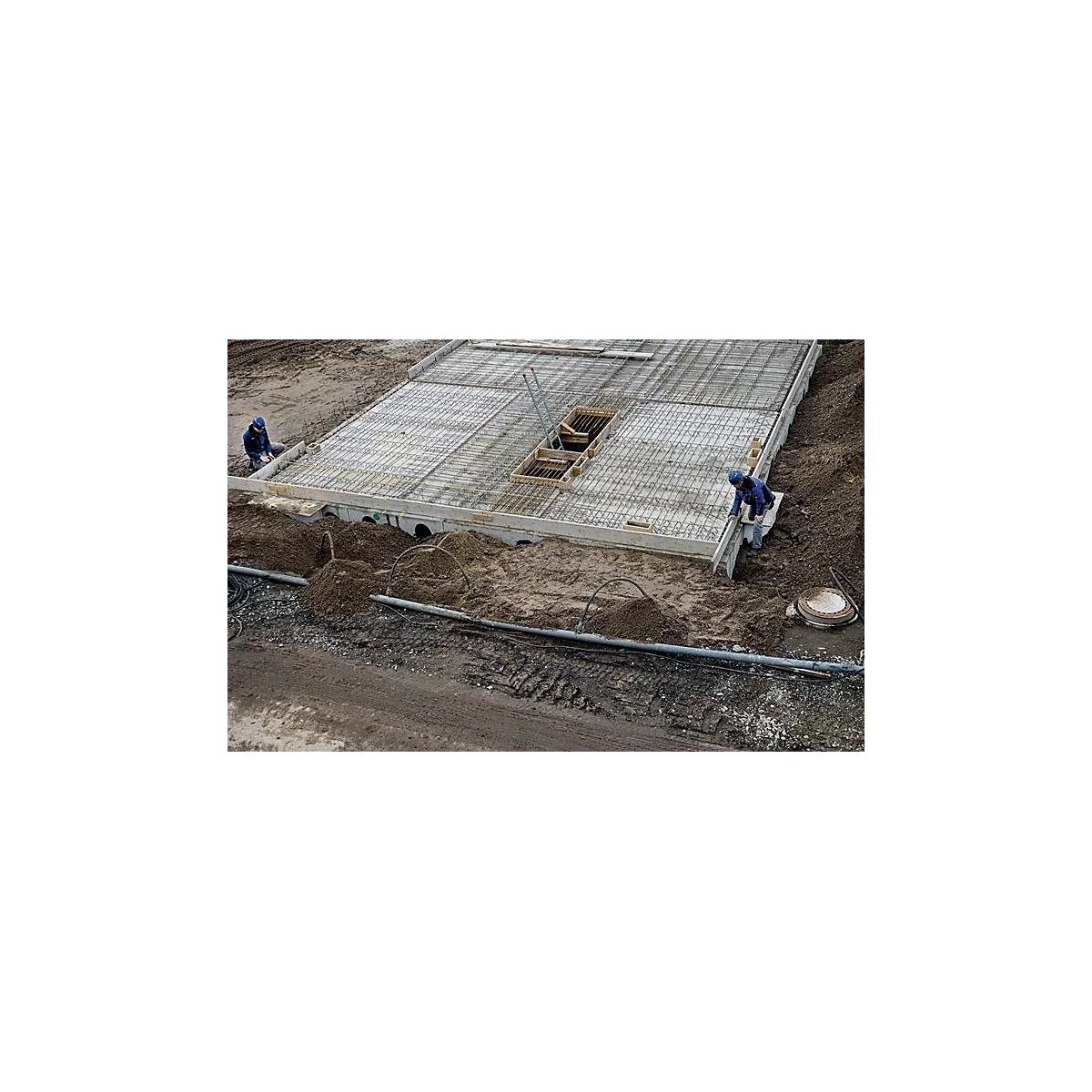 Bouwvakkers die werken aan een betonnen plaat. Staalmatten, aarde en pijpen zijn zichtbaar.