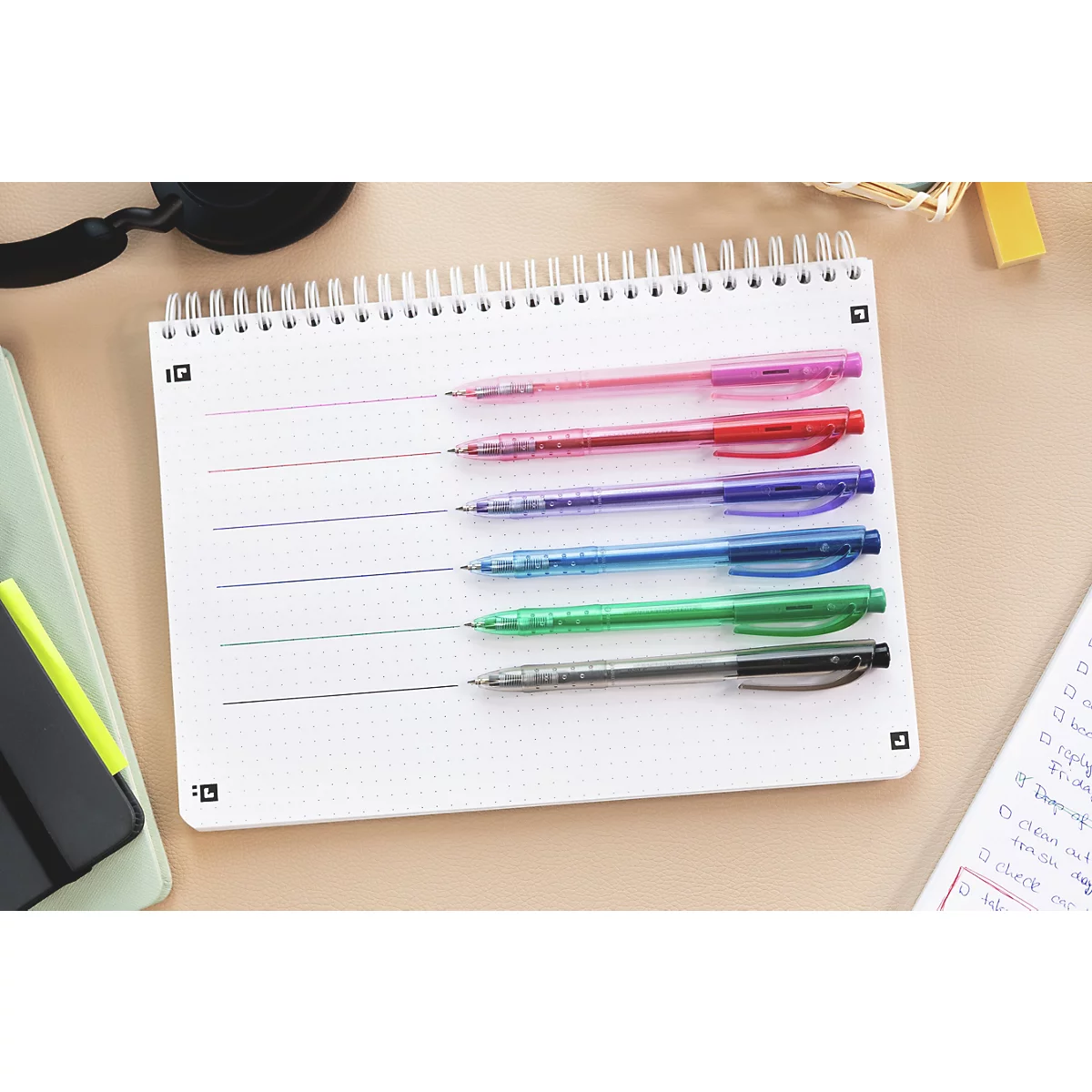 Mehrfarbige Stifte auf einem Notizblock mit verschiedenen Linien. Auf dem Tisch liegen weitere Schreibutensilien.