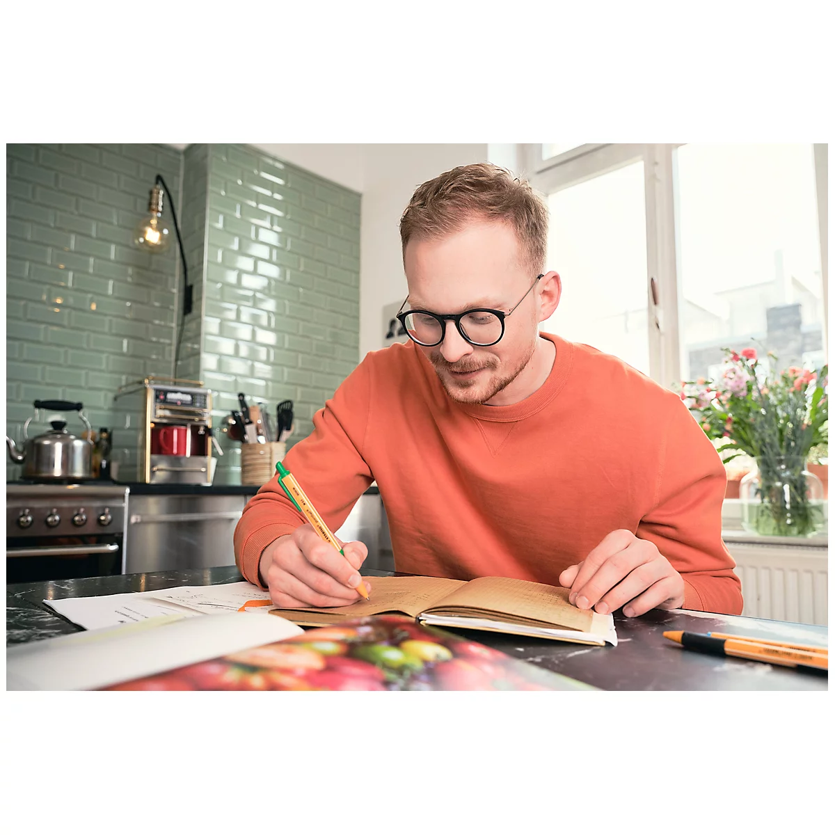 Ein Mann mit Brille und orangefarbenem Pullover sitzt und schreibt in einem Notizbuch. Im Hintergrund ist eine Küche zu sehen.