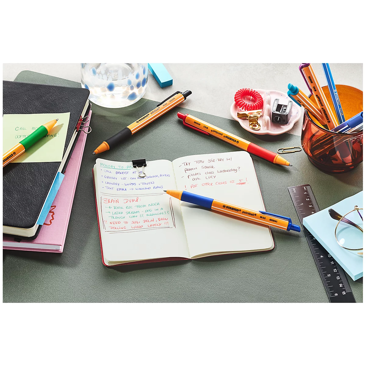 Nahaufnahme eines Schreibtischs mit Schreibwaren. Geöffnete Notizbücher mit handschriftlichen Notizen. Mehrere Stifte in verschiedenen Farben liegen herum.