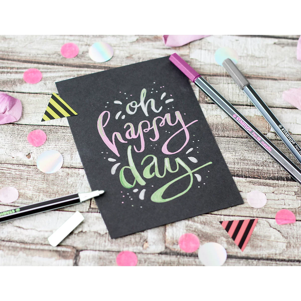 Carte noire avec "oh happy day" en lettres pastel. Confettis et stylos sur bois. Ambiance joyeuse !