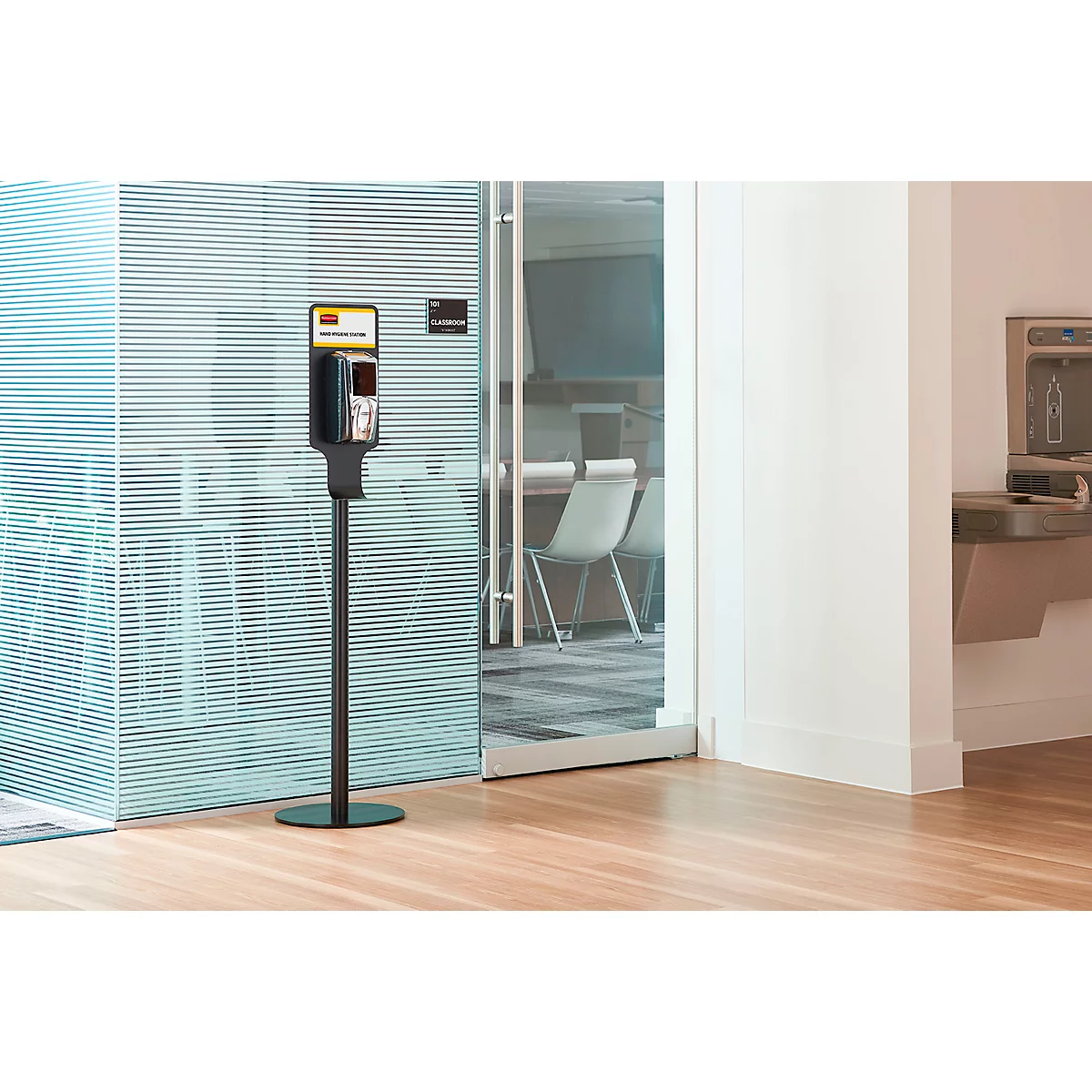 Ein schwarzer Desinfektionsmittelspender steht in einem hellen Raum. Hinter Glaswänden ist ein Konferenzraum zu sehen.