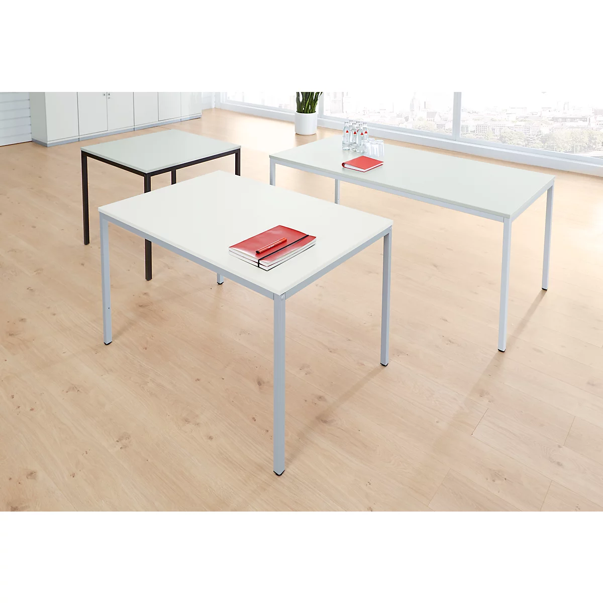 Drei Tische mit unterschiedlichen Formen. Auf dem mittigen Tisch liegt ein rotes Notizbuch. Holzfußboden.