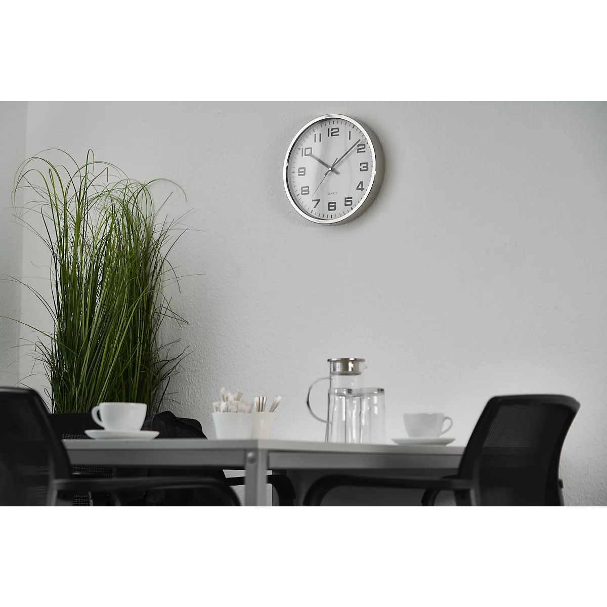 A table in a meeting room. Coffee cups, a glass jug and a vase with tall grass are on the table. A clock hangs on the wall.