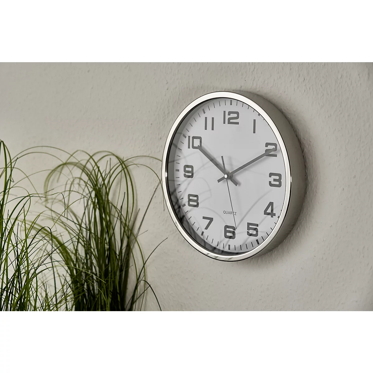 A round silver wall clock with black markings hangs on a light grey wall. Green plants are visible behind it.
