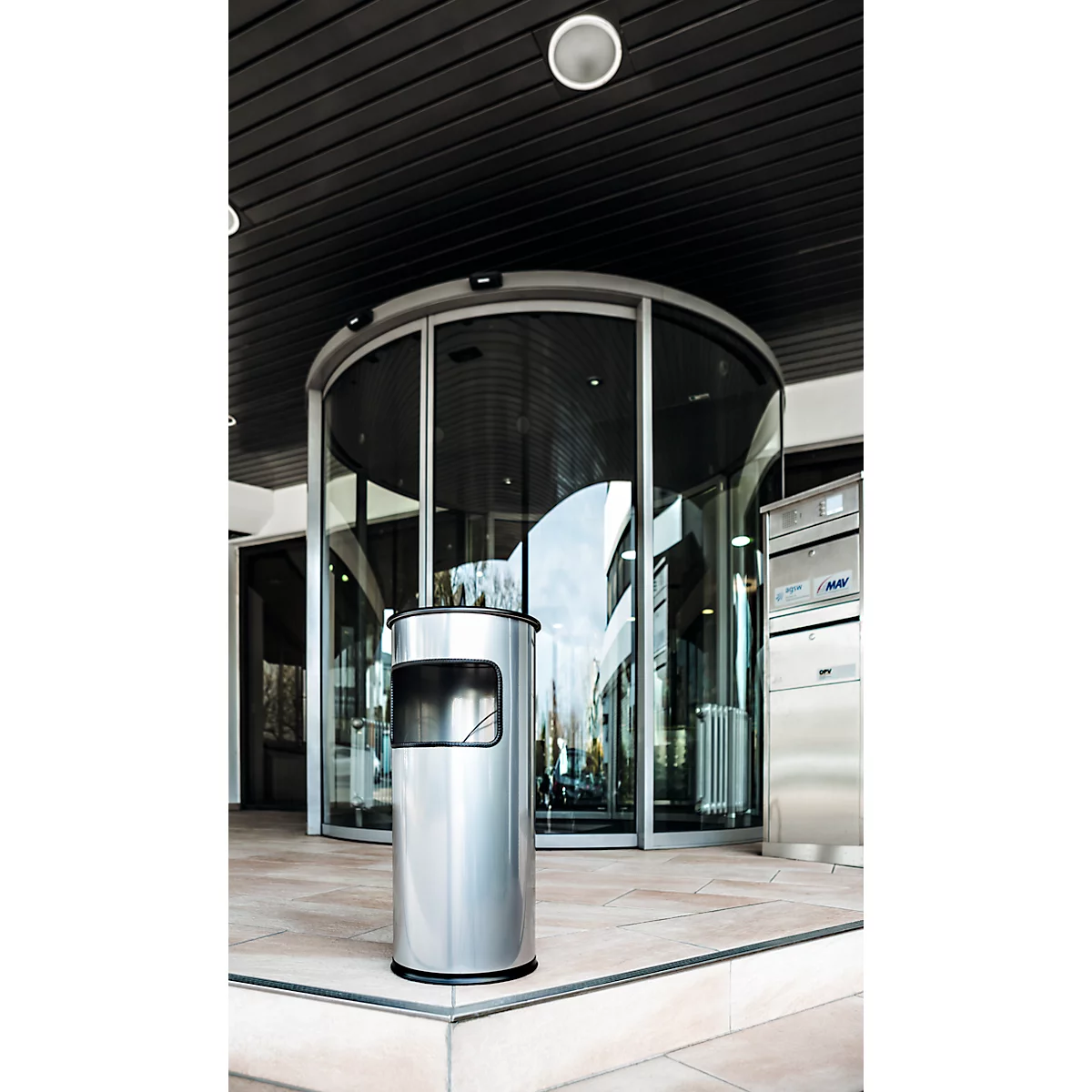 A cylindrical stainless steel waste bin stands in front of a glass revolving door at a modern entrance.