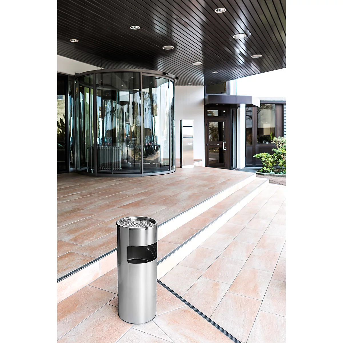 Stainless steel ashtray in front of an entrance hall. Revolving door, stairs and shiny ceiling in the background.