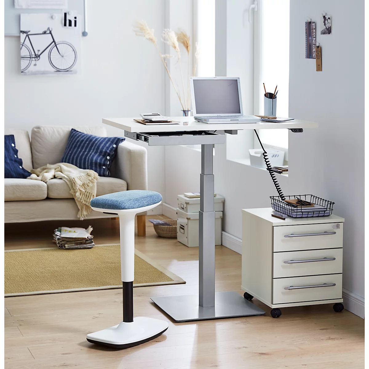 An adjustable height desk with a laptop, standing stool, drawer cabinet, and sofa in a bright room.
