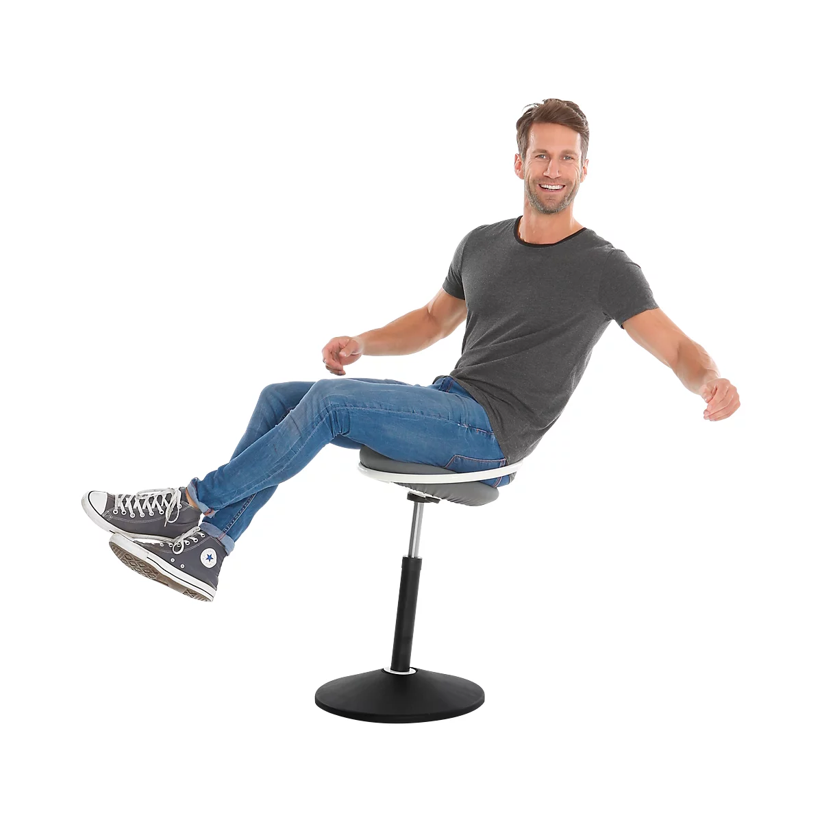 A man sits on a height-adjustable chair. He is smiling and has his arms outstretched. He is wearing jeans, a grey T-shirt and Chucks.