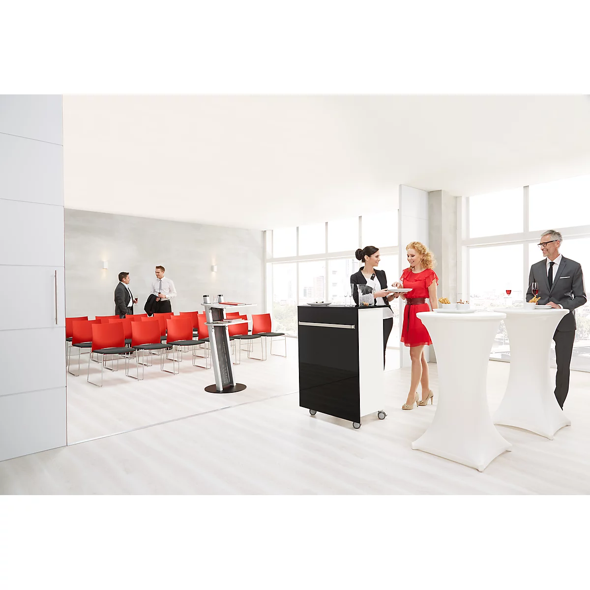 Several people are standing at high tables in a conference room. Red chairs are ready. Conference technology in the background.