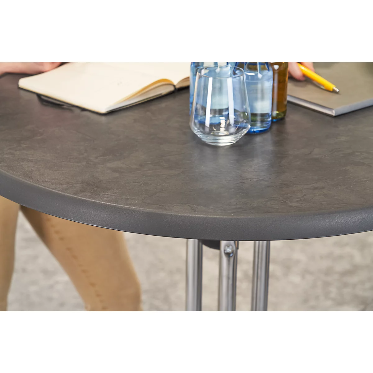 Round, anthracite-colored table. On it an open notebook, water bottles and glasses.