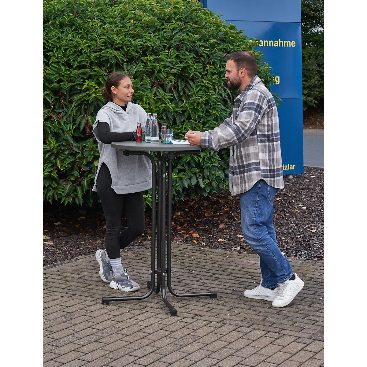 Two people stand at a high table. A woman in workout clothes and a man in a flannel shirt are talking.