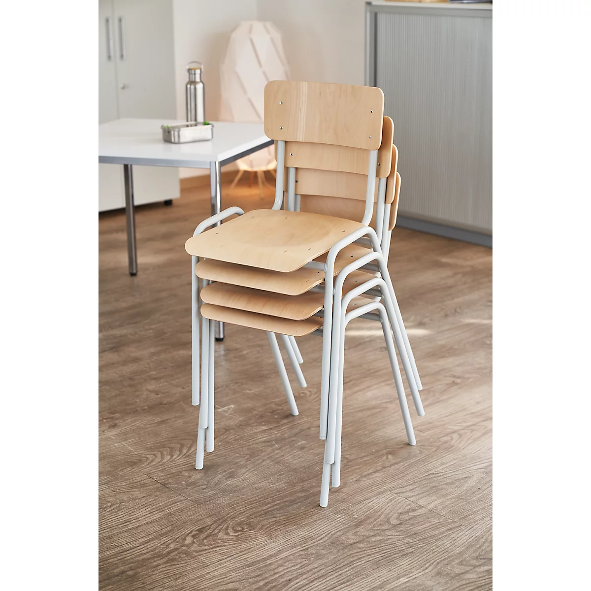 Gestapelde stoelen met licht hout en witte metalen frames. Op de achtergrond een tafel en een kast.