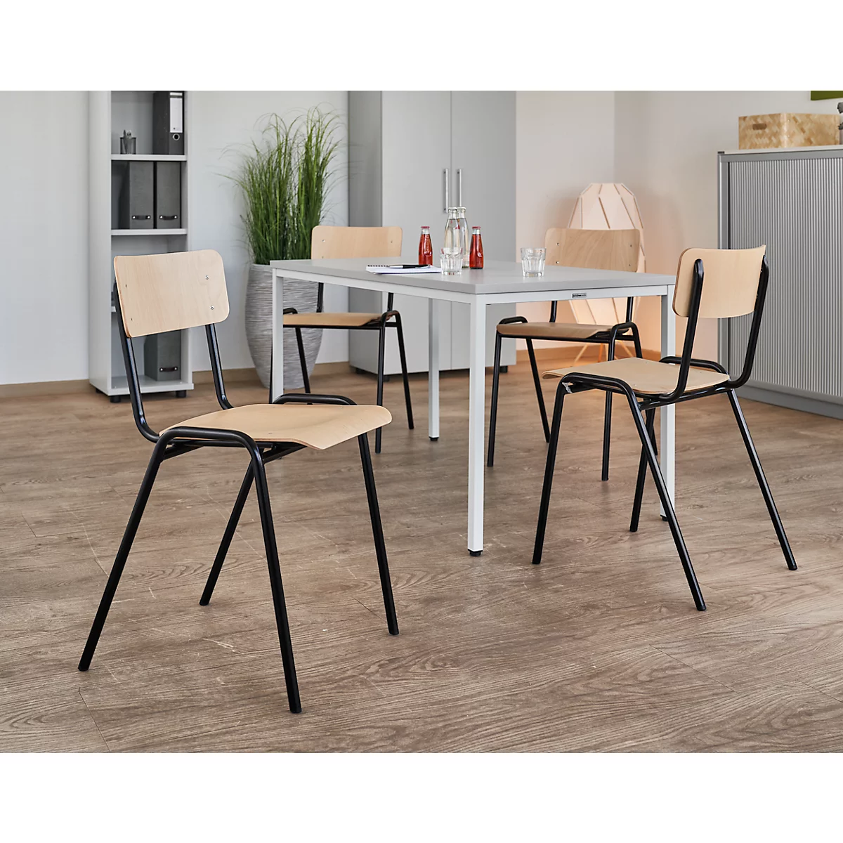 Een lichte houten tafel met vier stoelen. Zwarte metalen frames, lichtgekleurde zittingen. Rode en glazen flessen staan op tafel.