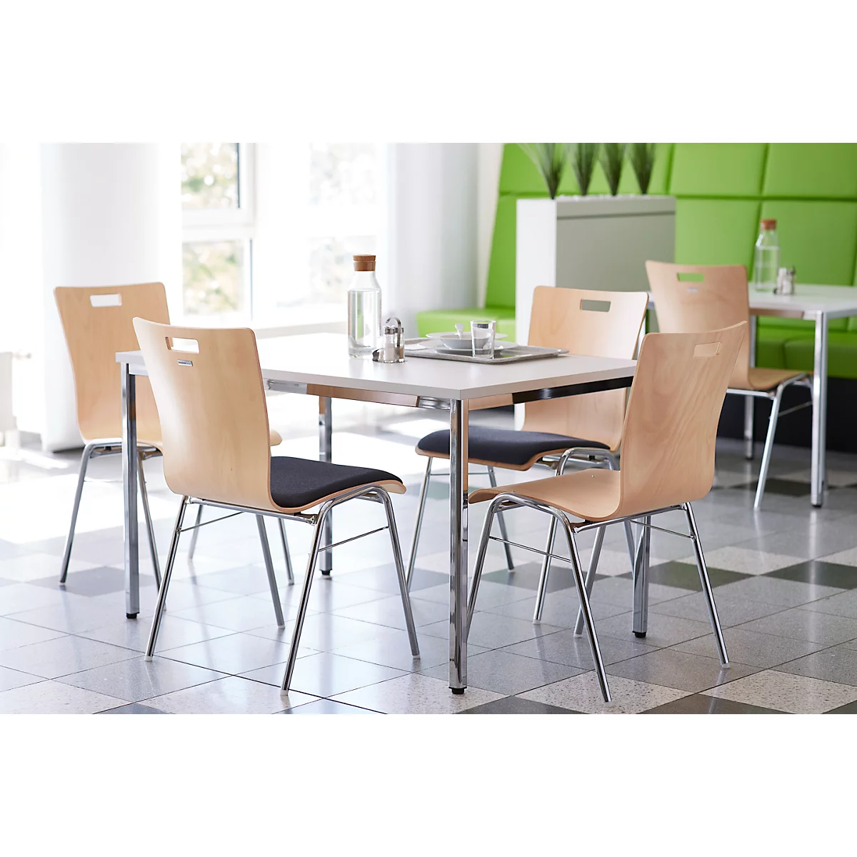 Een tafel met vier stoelen in een lichte kamer. De stoelen hebben houten rugleuningen en zwarte zittingen. Glazen en een fles staan op tafel.