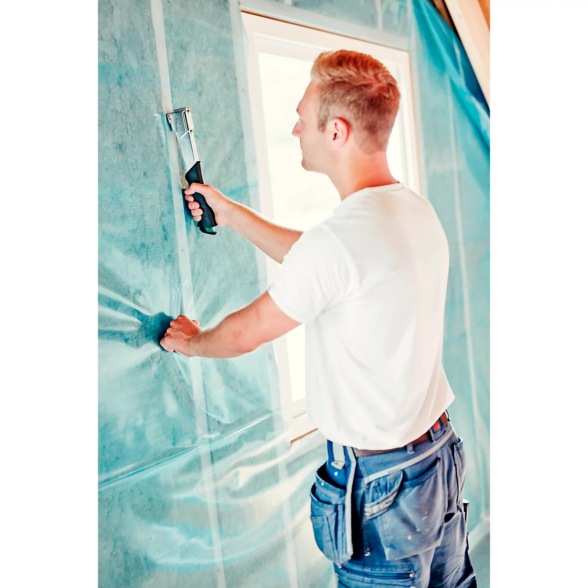 A man fastens foil to a wall with a stapler. He is wearing jeans and a white t-shirt.