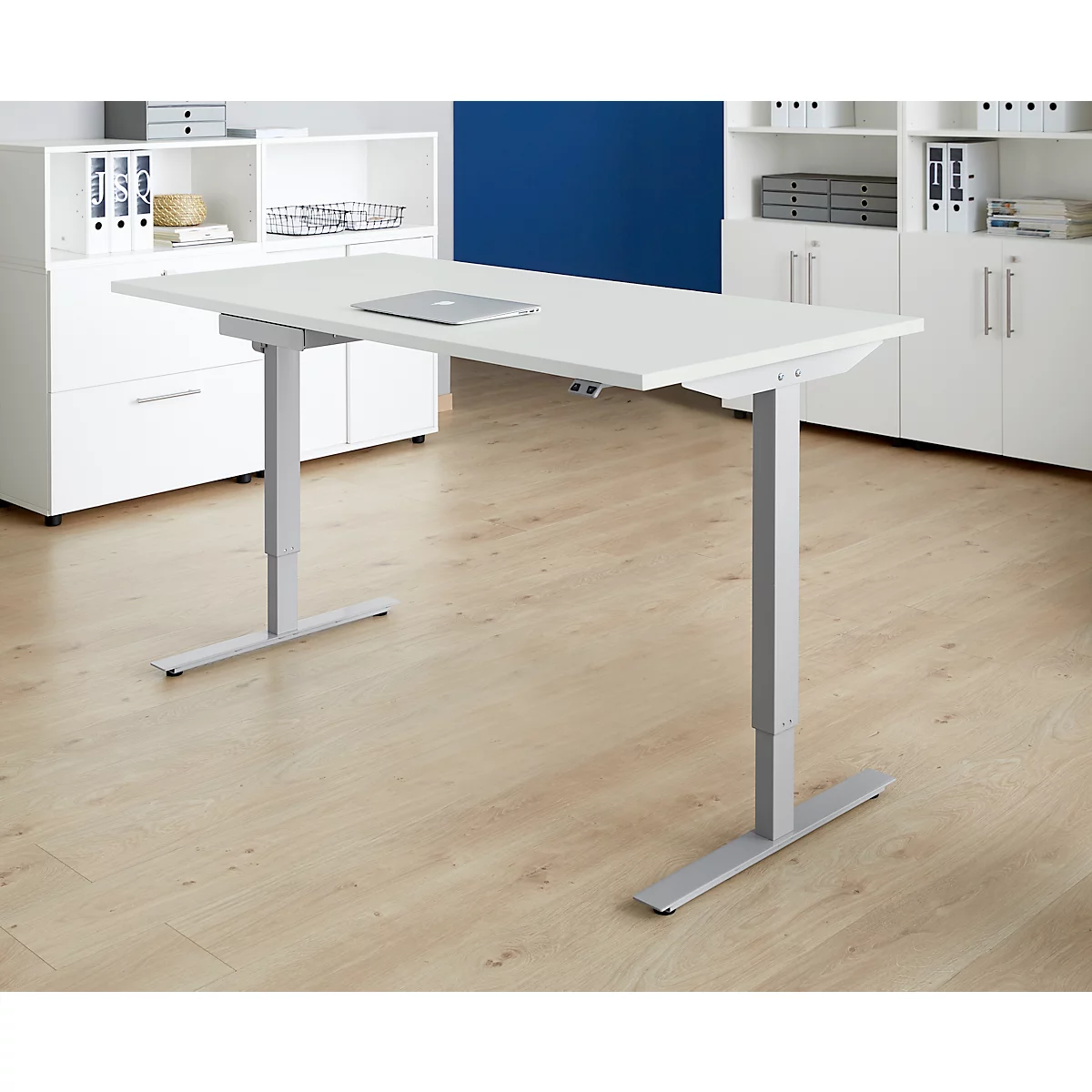 A white, height-adjustable desk with a laptop, set against a background of wooden flooring, white cabinets, and a blue wall.