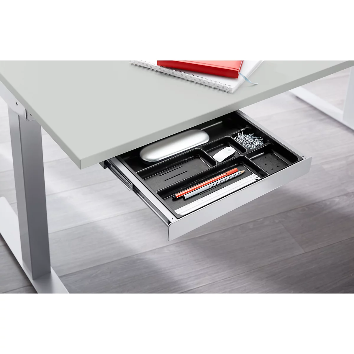 Close-up of a desk drawer with pens, mouse, paper clips. Desk top, grey, metallic legs.