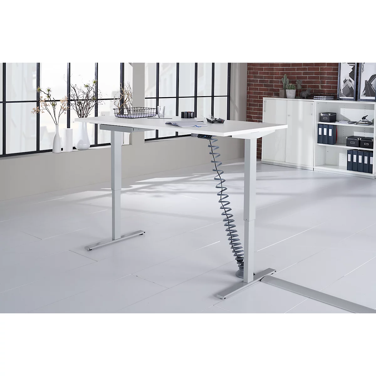 Height-adjustable desk. White tabletop, gray legs, behind it windows. Brick wall and cabinet in the background.
