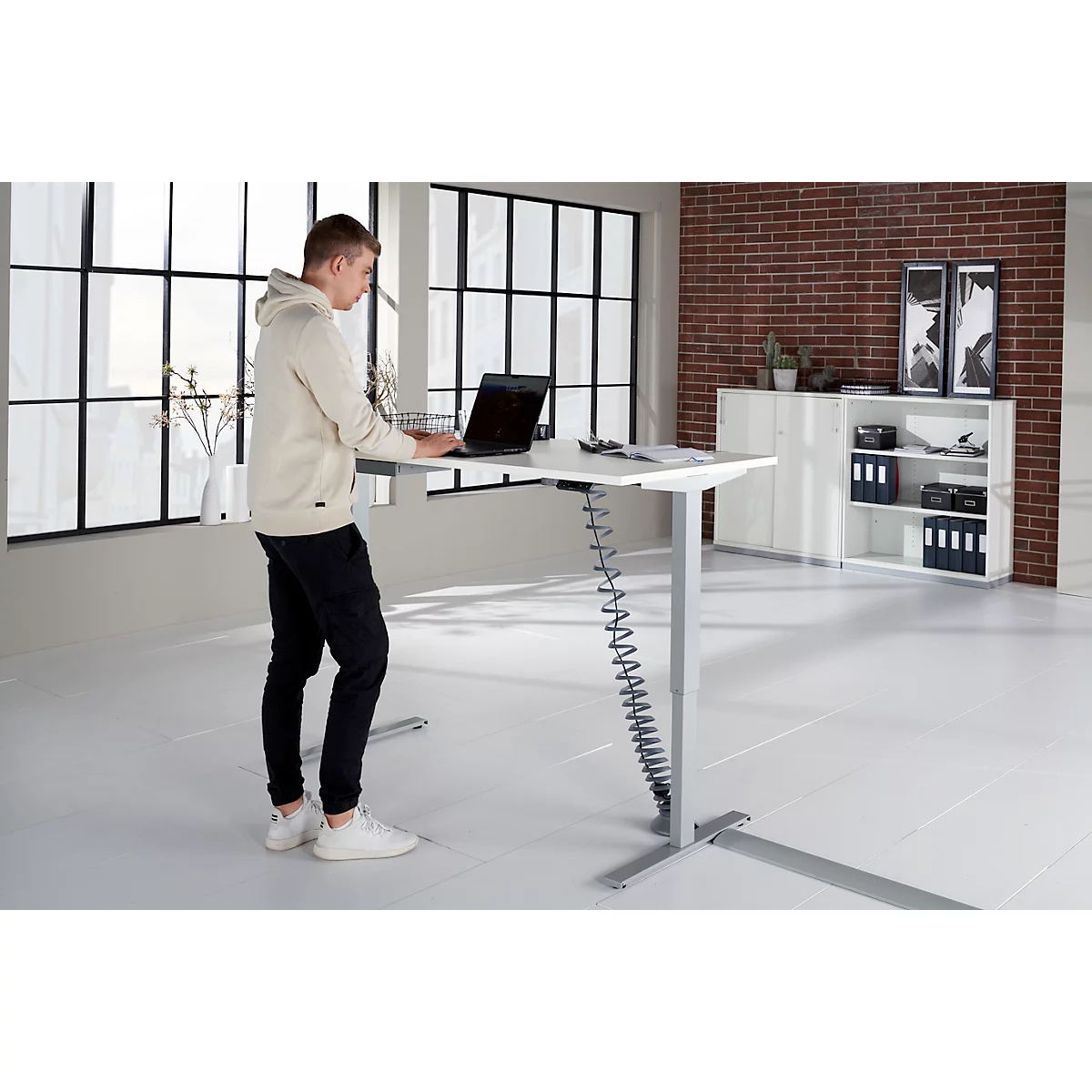 A man stands at a height-adjustable desk, working on a laptop, light clothing. A shelf and a window in the background.