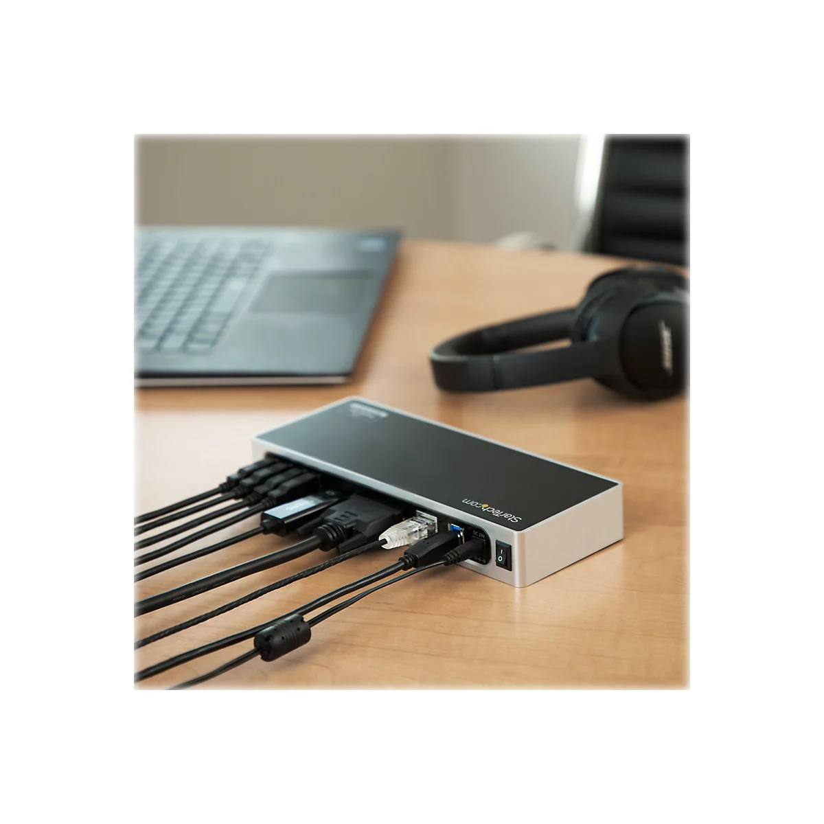 Ein schwarzer USB-C-Hub mit mehreren AnschlÃ¼ssen, an dem Kabel angeschlossen sind, liegt auf einem Holztisch neben einem Laptop und KopfhÃ¶rern.