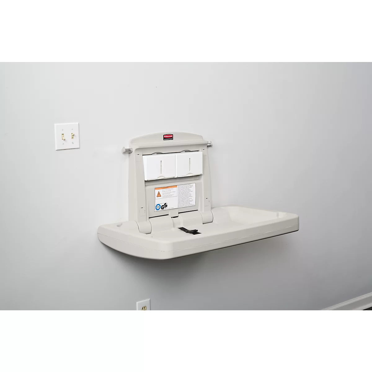 Table à langer murale gris clair, ouverte avec ceinture de sécurité, notice d'information. Prise électrique à gauche.