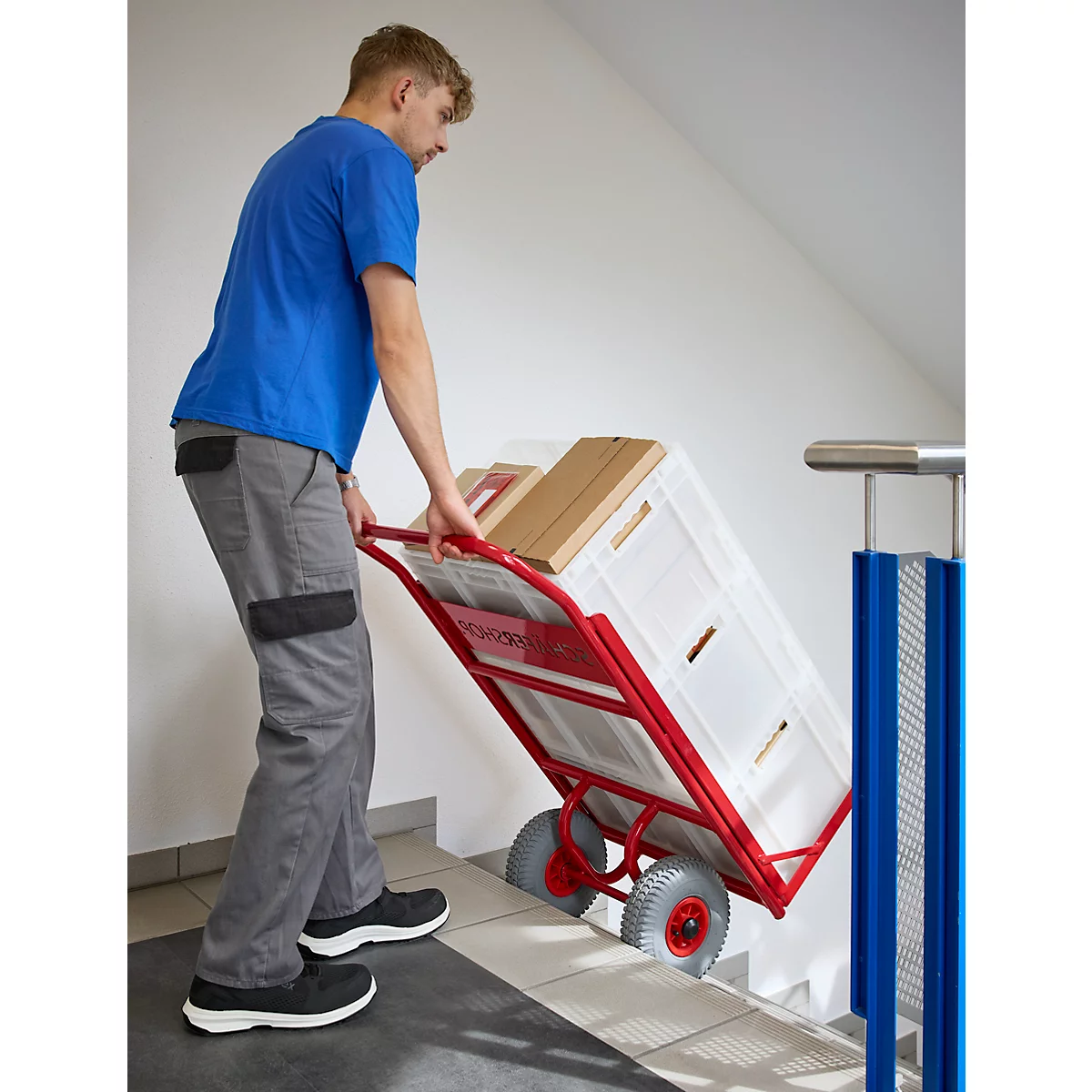 Een man in een blauw T-shirt trekt een beladen steekwagen een trap op. Op de kar staan een witte mand en dozen.