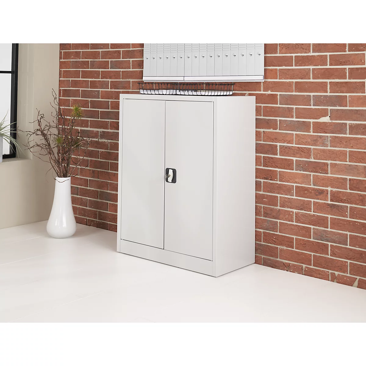 A grey steel cabinet with two doors in front of a brick wall. Folders are on the cabinet.
